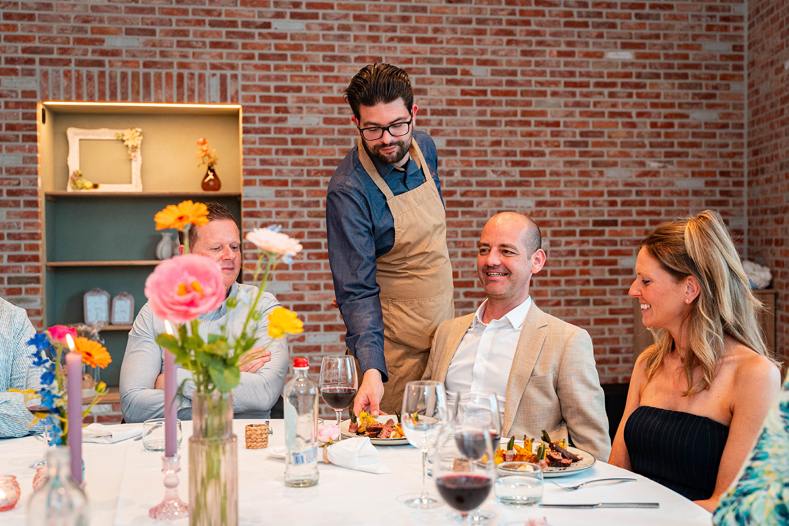 Chef Delivento serveert hoofdgerecht aan lachende gasten aan tafel met bloemen